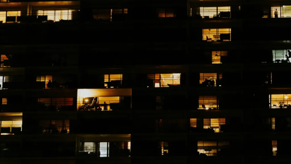 Large apartment building exterior at night showing a mix of dark empty units and brightly lit occupied windows.