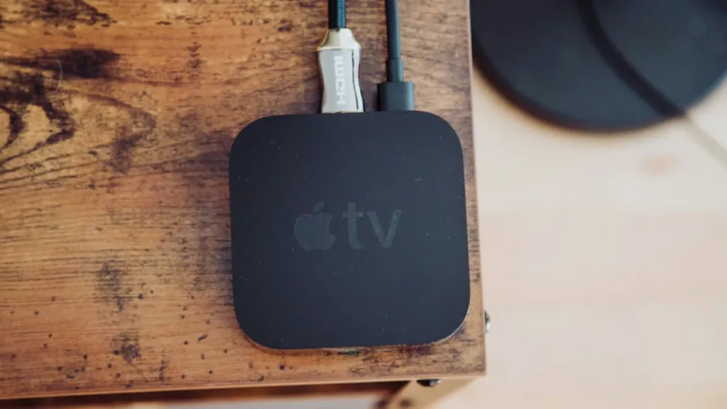 Apple TV 4K media box connected with a braided HDMI cable on a wooden TV stand.