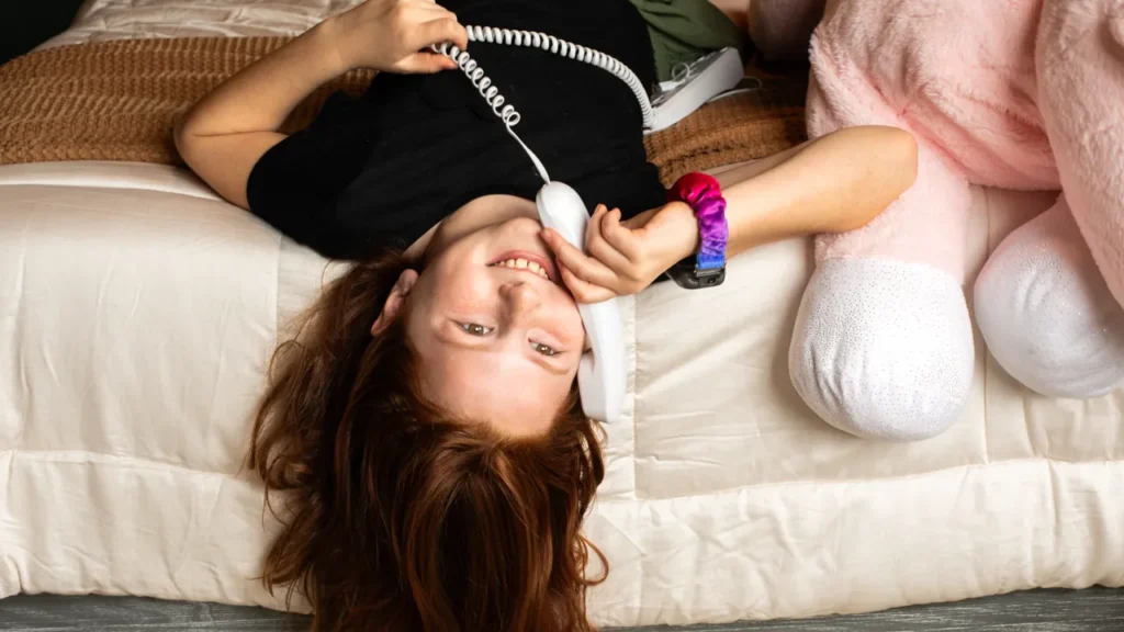 A smiling young girl laying upside down on a bed while talking on a white kid-safe Pinwheel Home.