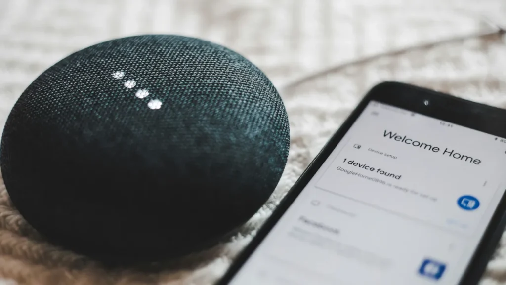 A Google Home Mini speaker sitting next to a smartphone displaying the "Welcome Home" device setup screen.