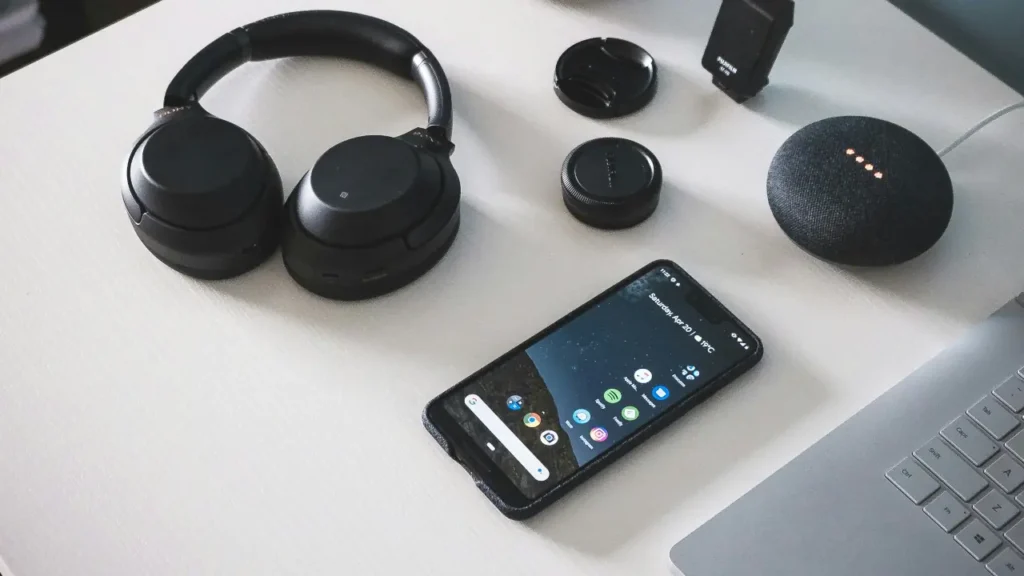 A smartphone, black wireless headphones, and a glowing smart speaker arranged on a clean white desk.
