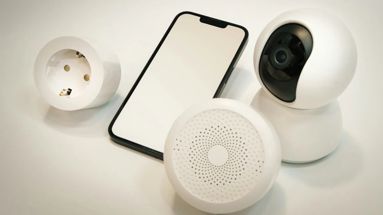 Smartphone lying next to a white smart plug, a home security camera and a smart hub on a white table